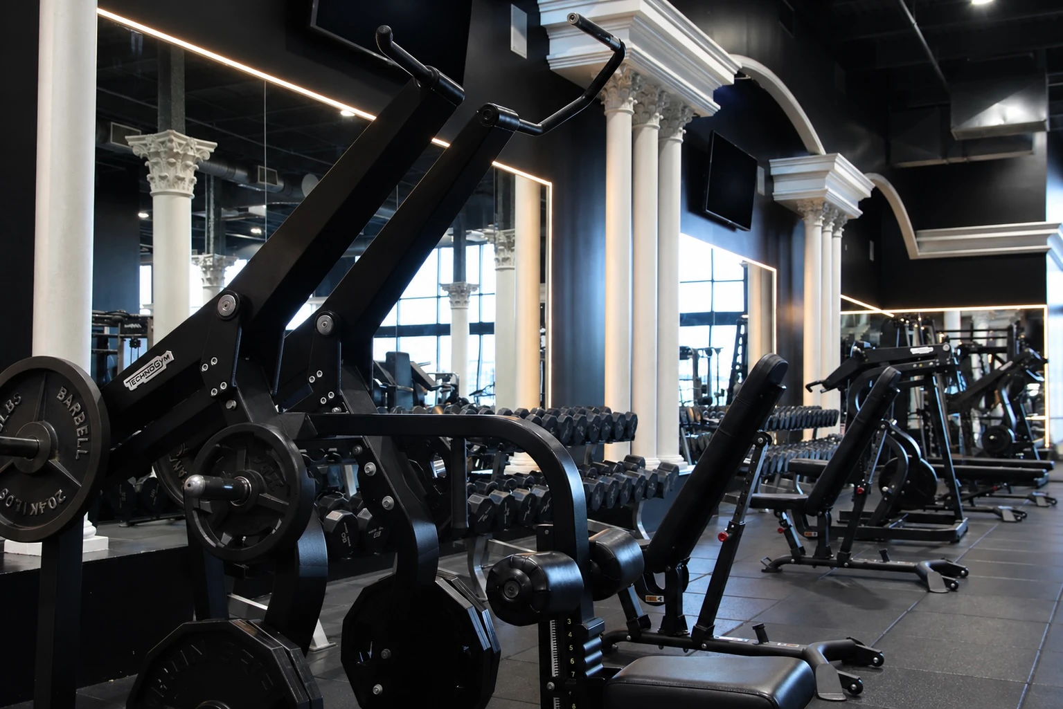 Plate-loaded machines and dumbbell racks between white columns at XCEL Athletic Lifestyle, a rooftop gym in Hoboken, NJ