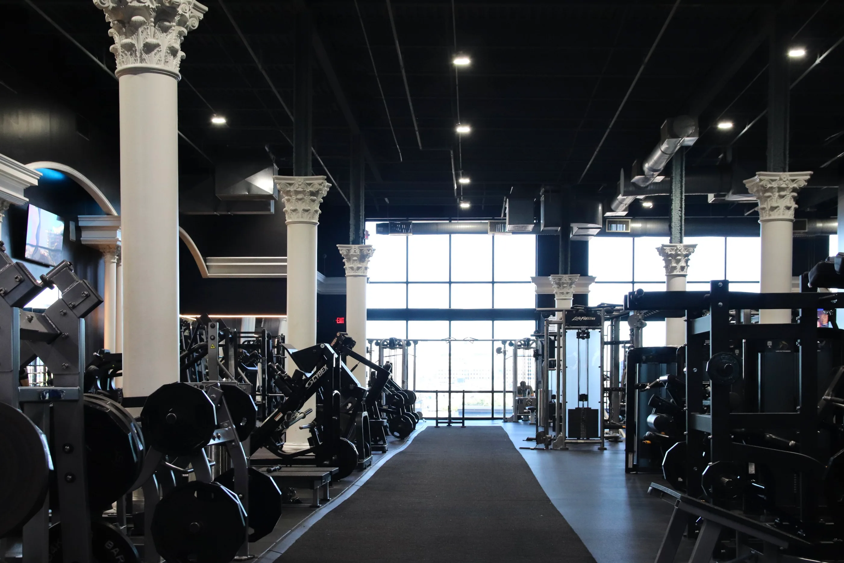 Strength machines between white columns and floor-to-ceiling windows at XCEL Athletic Lifestyle, a rooftop gym in Hoboken, NJ
